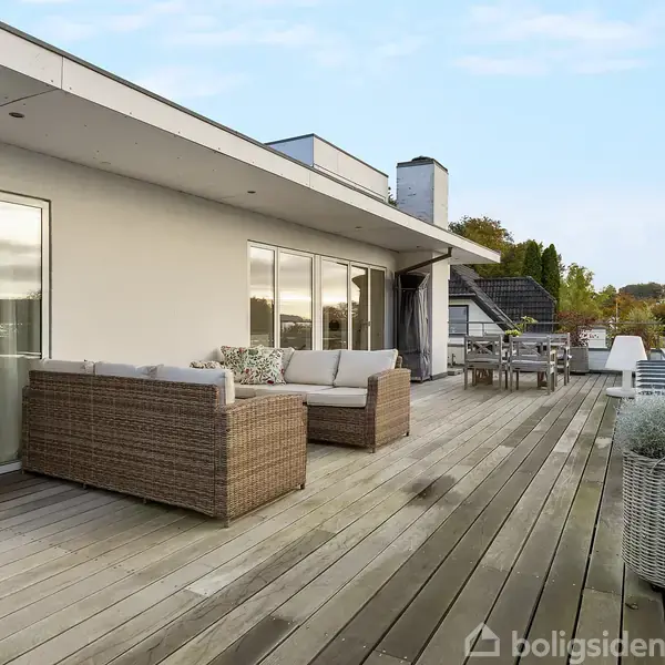 Rattan sofas står på en stor træterrasse med glasrækværk, gryder med planter og udsigt over trætoppe. En stige fører til en højere platform.