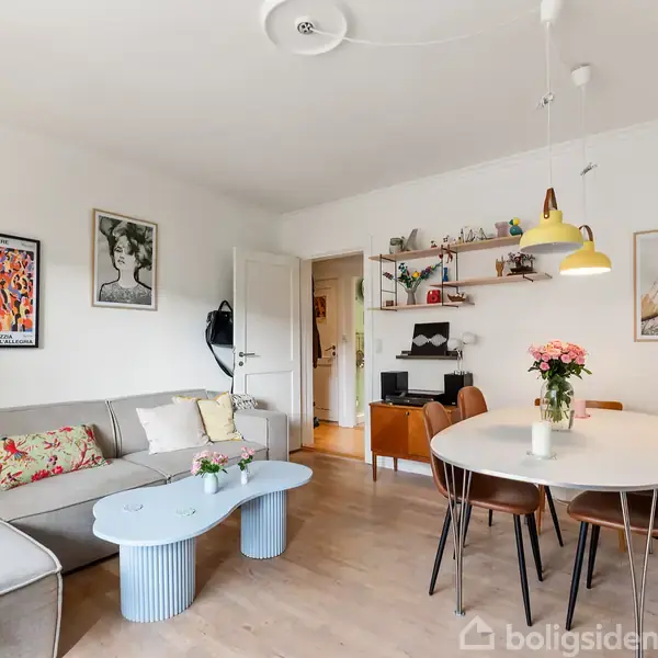En stue med gråt hjørnesofa og et lyseblåt sofabord. Et rundt spisebord med fire stole og en vase med blomster. Lys fra en gul lampe og kunst på væggene.