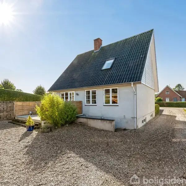 Et hvidt hus med sort tag står stille på en grusbelagt grund. Solen skinner fra en klar himmel, omgivet af andre boliger og grøn beplantning.
