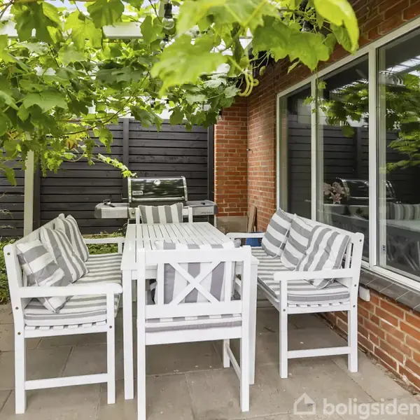 Hvide havemøbler med grå- og hvidstribede puder står under en pergola med grønne blade. En grill ses ved en mørk trævæg, samt et stort vindue i baggrunden.