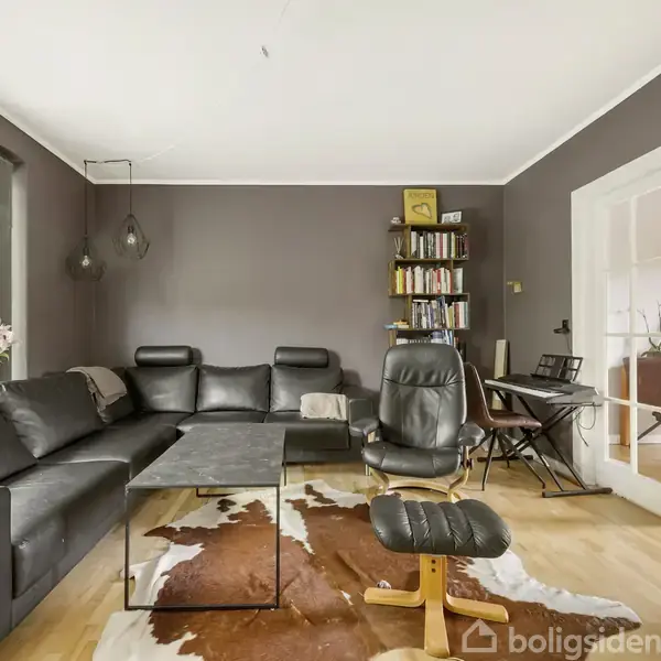 Sort lædersofa og stol står i en stue, omgivet af et sofabord på et brunt og hvidt tæppe. En reol fyldt med bøger står i hjørnet, og store vinduer giver udsigt til en terrasse.