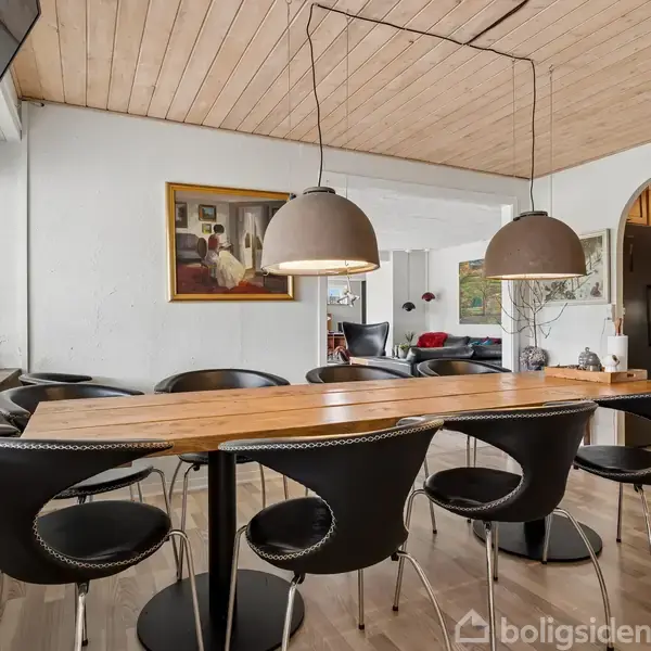 Lang træbord omgivet af sorte stole står i spisestue med træloft. To store pendellamper hænger over bordet. I baggrunden ses en sofa og maleri på væggen.