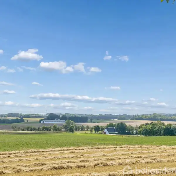 Grønt landskab med høstede marker, strøer af halm i forgrunden; bagved ses spredte træer og lader mod en blå himmel med lette skyer.
