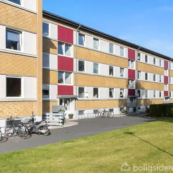 En række etagebygninger med gule mursten og røde paneler står langs en asfaltvej. Foran bygningerne er flere parkerede cykler. En velplejet græsplæne ligger i forgrunden.