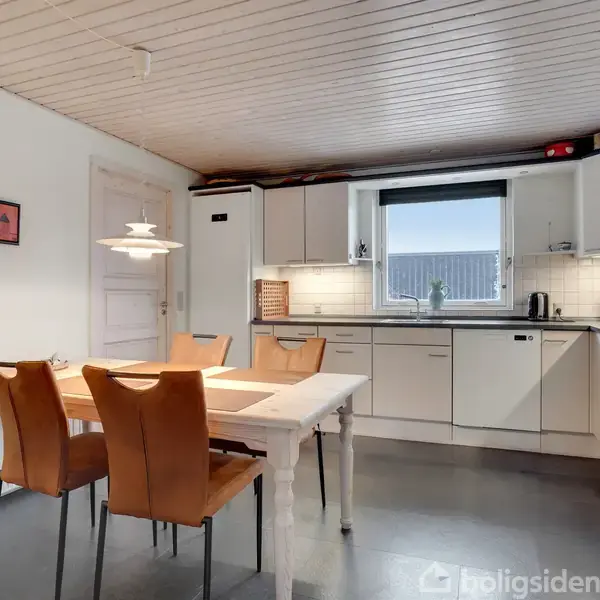 Et køkken med hvide skabe og en kogeplade. Et hvidt bord er omgivet af fire brune stole. En lampe hænger over bordet, og en enkeltstående dør er ved siden af.