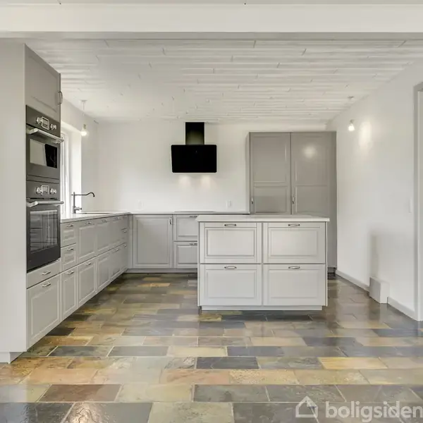 Køkkenet har grå skabe og en central køkkenø. En integreret ovn ses til venstre, og en emhætte hænger over komfuret. Flisegulvet er flerfarvet, og rummet er lyst.