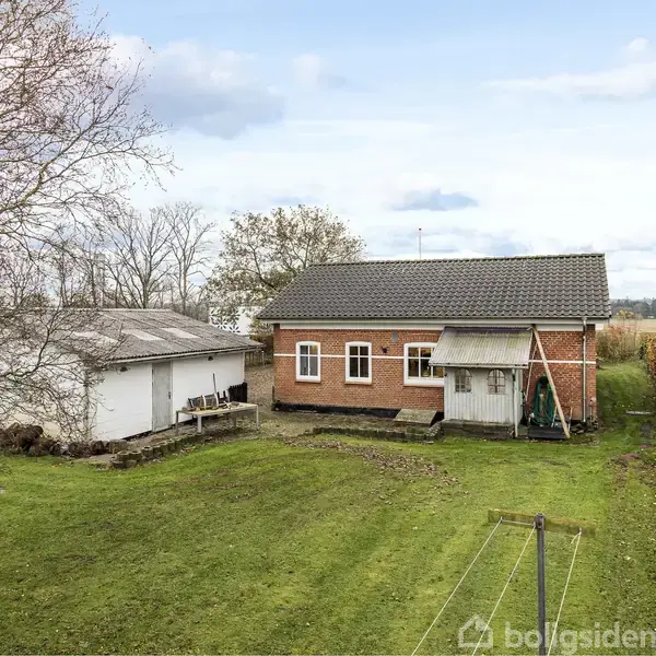 Rødt murstenshus med gråt tag står i en grøn have ved siden af en hvid bygning. Træer og åben mark omgiver området under en blå, skydækket himmel.