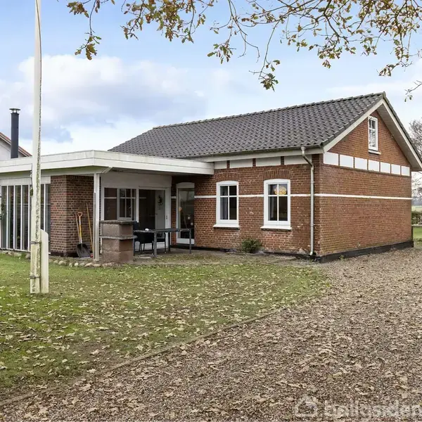 Et rødt murstenshus med sort tag ligger ved siden af en grusvej. Foran er en græsplæne og et flagstang. En garage ses til højre.