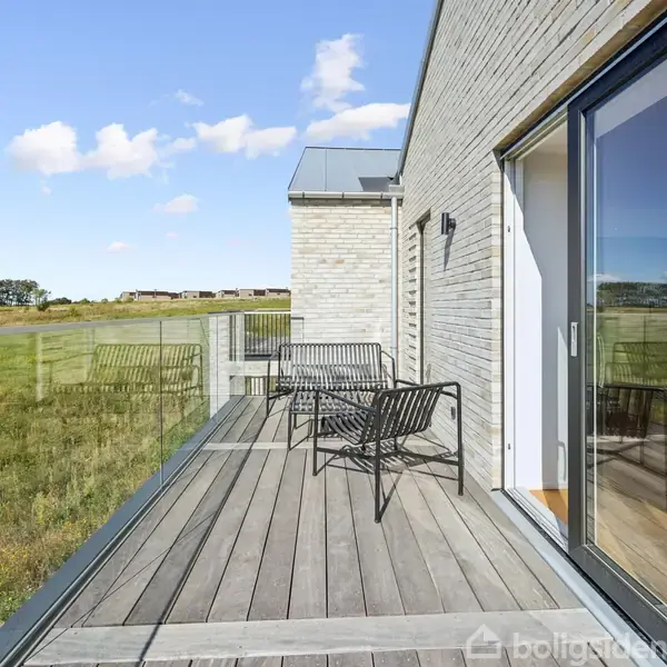 En træterrasse med to sorte stole står langs en murstensbygning. Terrassen har glasrækværk og udsigt til en stor grøn mark og en række træer i baggrunden.