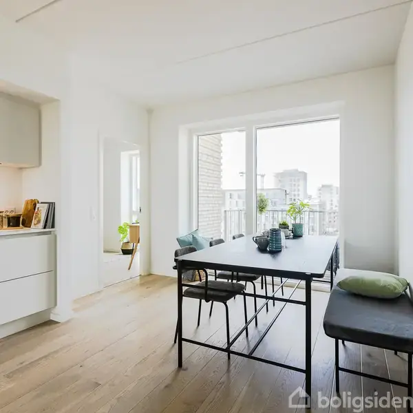 Et moderne køkken-alrum med et sort spisebord omgivet af stole og en bænk, ved siden af store vinduer, der giver udsigt til bybygninger. Køkkenet er til venstre med hvide skabe.