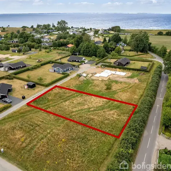 Græsareal indrammet af rød linje ligger i et villakvarter nær en vej. Baggrunden viser flere huse, træer og havudsigt i det fjerne.