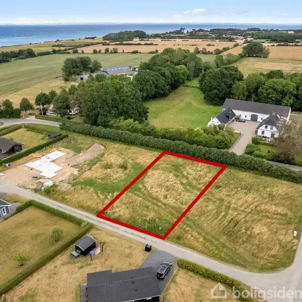 Tomt omkranset af rød linje ligger blandt åbne marker og spredte huse i et landligt område nær kysten. Inkluderer veje og grønne områder, med havet i baggrunden.