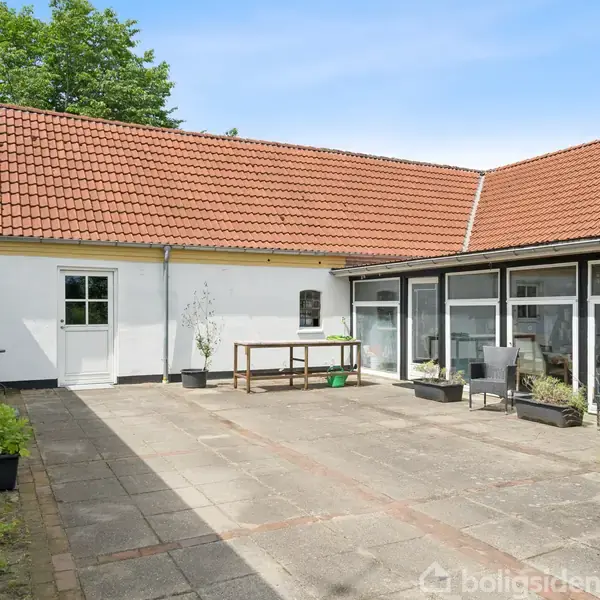 Indre gårdhave med flisebelægning omgivet af hvidmalede bygninger med rødt tegltag. Terrassemøbler og krukker med planter står rundt omkring i gården.