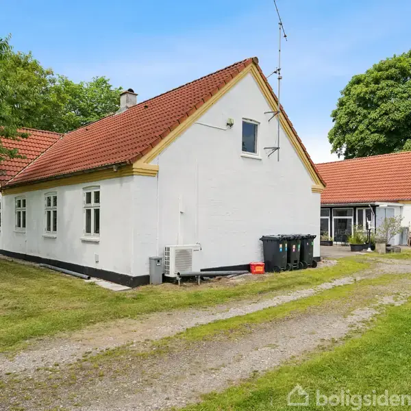 Hvidt hus med rødt tegltag, roligt beliggende omgivet af grønne træer og græs, grussti fører til indgang.