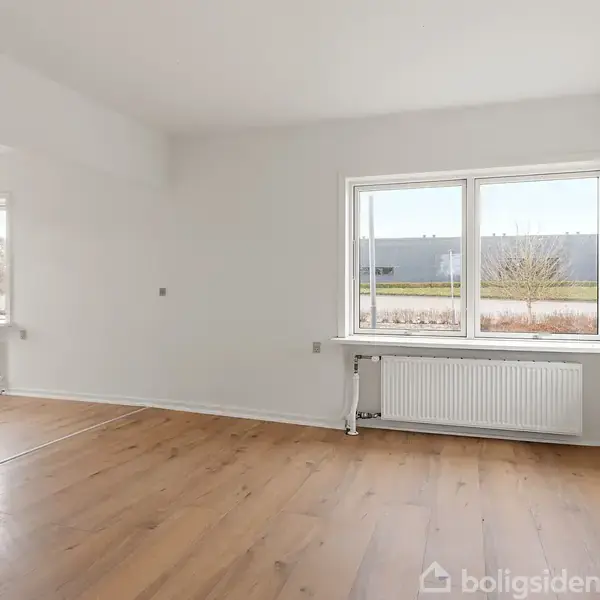 Rummeligt, lyst værelse med lyse trægulve og hvide vægge. Store vinduer mod en flad, udendørs scene udenfor. Radiator placeret under vinduerne.