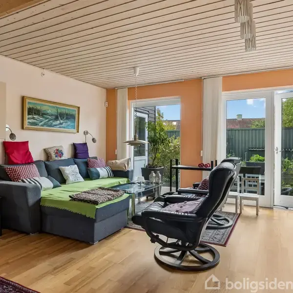 Stue med sort sofa og røde puder, to lænestole, sofabord med glas, maleri på væggen, store glasdøre fører til terrasse og have med grønne planter.