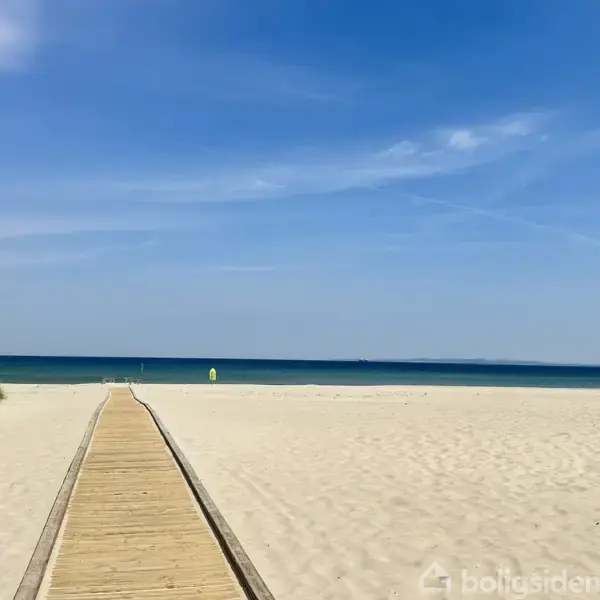 En træsti strækker sig over en bred sandstrand mod det blå hav. Himlen er klar og blå, og der er lidt vegetation til venstre.