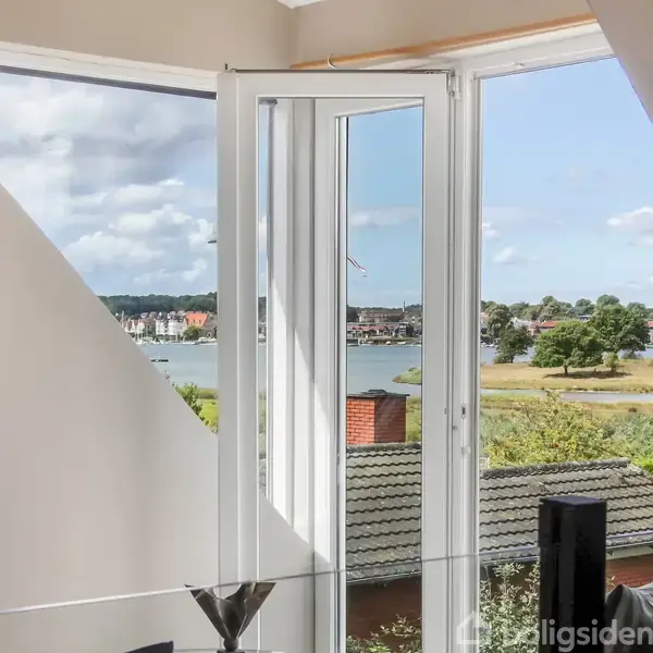 Et åbent vindue viser en malerisk udsigt over en sø med trækroner, marker og huse på den anden side. Rummet indeholder et maleri på væggen og en glasvase.