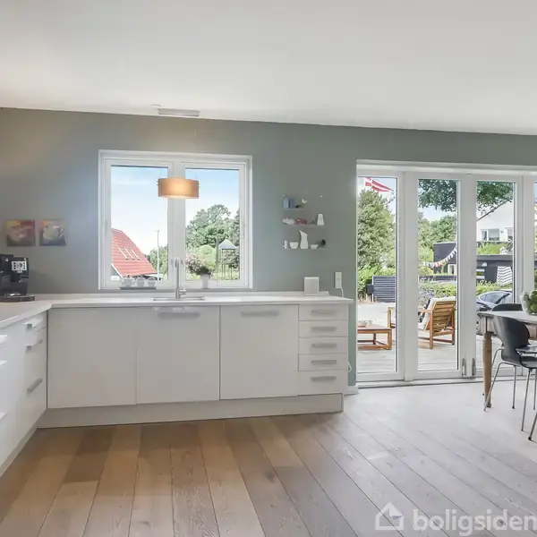 Et lyst køkken med hvide skabe og træbord. Store vinduer åbner mod en terrasse med havemøbler. Grønne omgivelser er synlige udenfor gennem de åbne døre.
