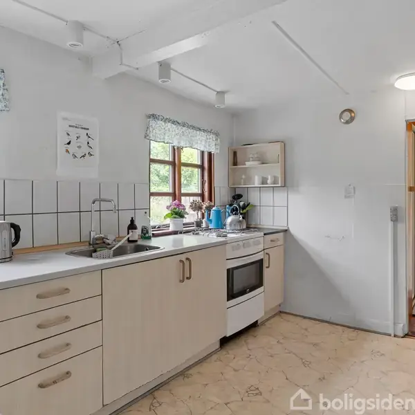 Køkken med beige skabe, komfur og vask; rolig handling med organiserede køkkenredskaber. Sollys gennem vinduer med blomster. Lyst, rummeligt miljø skaber en hyggelig atmosfære.