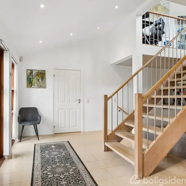 En entre med en trappe af træ, der fører opad. Ved siden af trappen er der en grå stol og en dør. Et tæppe er placeret på gulvet.