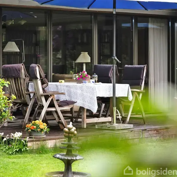 En terrasse med træbord og stole under parasol. Bordet har hvid dug, blomster og lys. Haven omkring er grøn med blomster og en lille springvand.