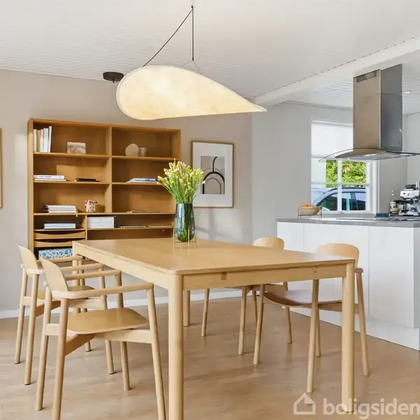 En træspisebord med fire stole står i et lyst køkken-alrum. En moderne loftlampe hænger over bordet. Bagerst er en reol med dekorationer og billeder på væggen.