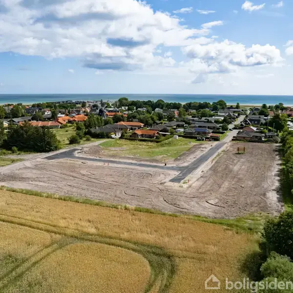 En stor byggeplads ligger stille i et forstadsområde nær kysten med marker, huse og campingpladser omkring, under en delvis skyet himmel. Ingen tekst er til stede.