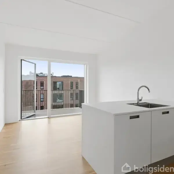 Hvid køkkenø med vask står i et lyst rum med trægulv. En åben dør fører ud til en balkon med udsigt til en murstensbygning.