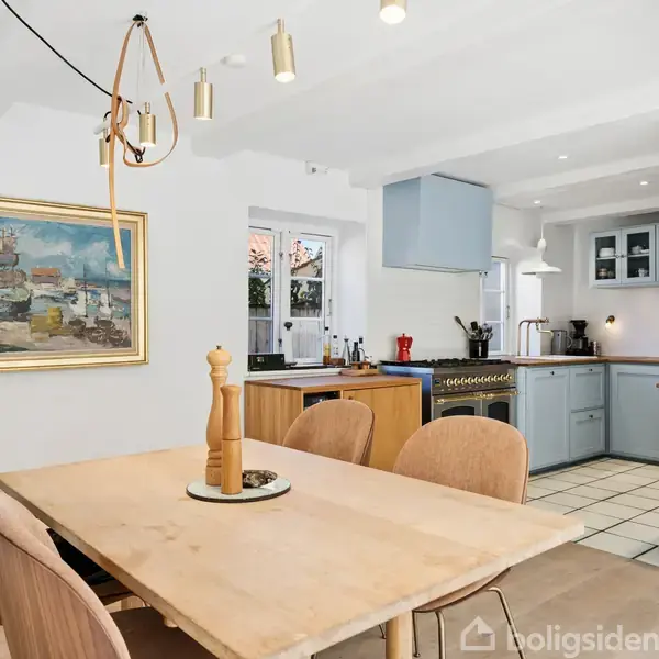 Bord med peberkværn, omgivet af fire stole, placeret i en spiseplads. I baggrunden ses et køkken med himmelblå skabsfronter, flisegulv og et vindue med udsigt til en gårdhave.