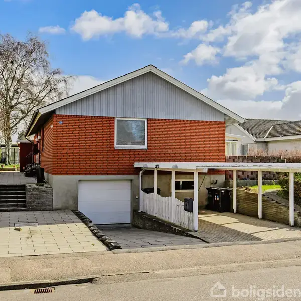 Et rødt murstenshus med en carport ved siden af, omkranset af en velplejet have i et forstadsområde. En skyfuld himmel danner baggrunden for stedet.