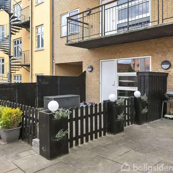 En udendørs terrasse med sorte stakitter og plantede blomster står nær en bygning med gule vægge. Trappen i baggrunden fører til øvre etager. Grill og planter ses på terrassen.