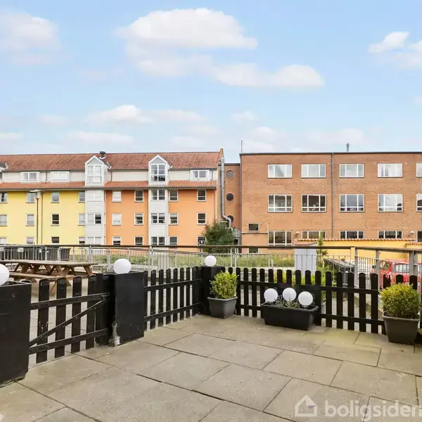 En terrasse med sorte hegn og store fliser har planter i krukker og dekorative lamper. I baggrunden ses flerfarvede lejlighedsbygninger under en let skyet himmel.