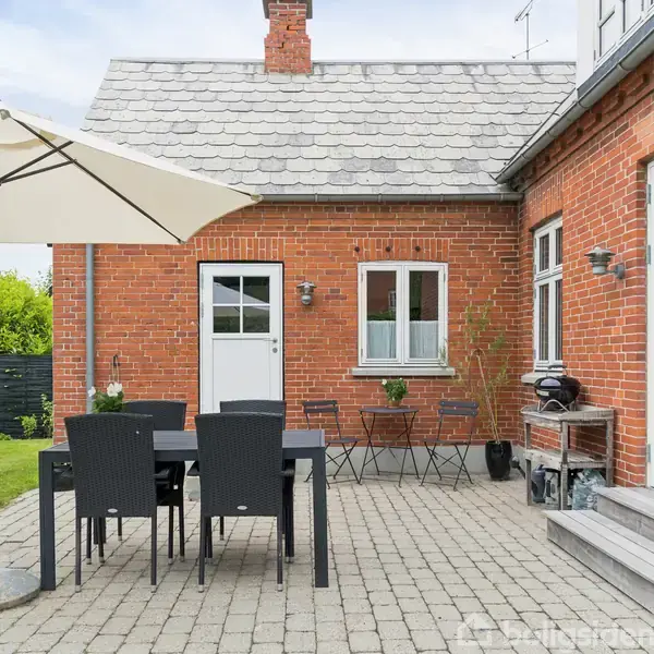 En terrasse med fliser viser et bord og fem stole under en stor parasol. Omgivet af et rødt murstenshus og en grøn have i baggrunden.