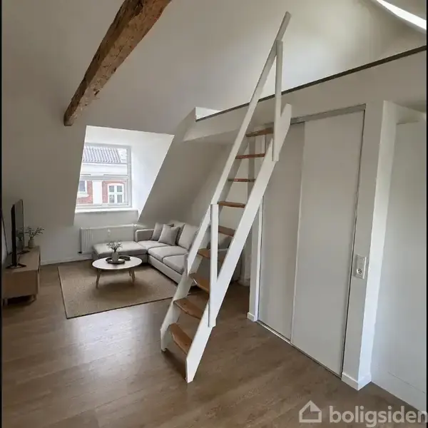 En stige fører til en hems i en moderne stue med et skråvindue. Rummet indeholder en sofa, et lille rundt bord og en TV-enhed.