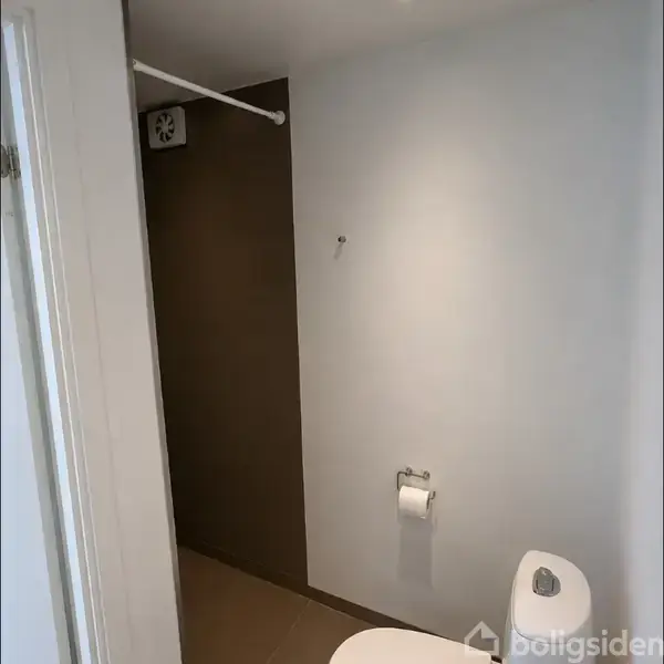 Et lille badeværelse viser en brun væg med en ventilation og en papirholder med toiletpapir. En hvid dørkarm ses til venstre, hvilket indikerer et rumindgang.