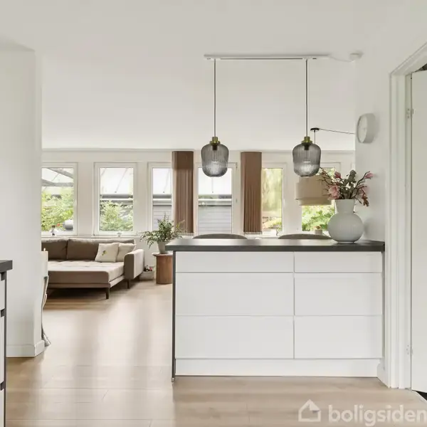 Køkken med hvide skabe, sort bordplade og hængelamper. Stue med sofa i baggrunden, store vinduer og lys trælaminatgulv skaber en lys og moderne atmosfære.