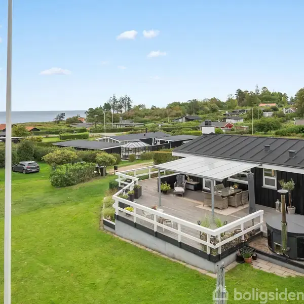 Sort sommerhus med terrasse, havemøbler og spabad, beliggende i en grønt område med græsplæne. Kysten og havet ses i baggrunden, omgivet af flere huse og træer.