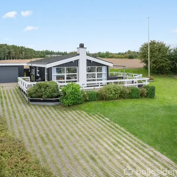 Et sort-hvidt hus med en hvid terrasse står i en velplejet have med græs og buske. Foran er der en belagt indkørsel. I baggrunden ses en mark og skov.