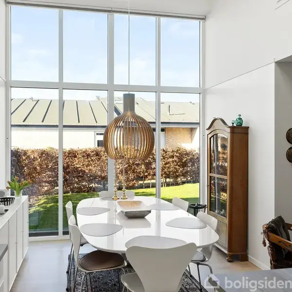 Spisebord med moderne stole står under en stor, rund lampe i et lyst rum. Store vinduer viser en have med hække udenfor. Vægkunst ses til venstre, skab til højre.