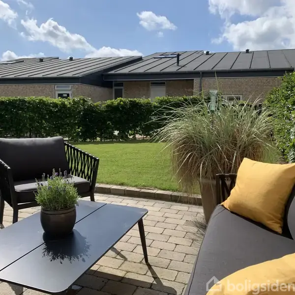 Sort udendørs lounge møblement står på en terrasse med stenbelægning, dekoreret med en plante på bordet og gule puder på sofaen. Haven omkranses af en hæk, under en blå himmel.