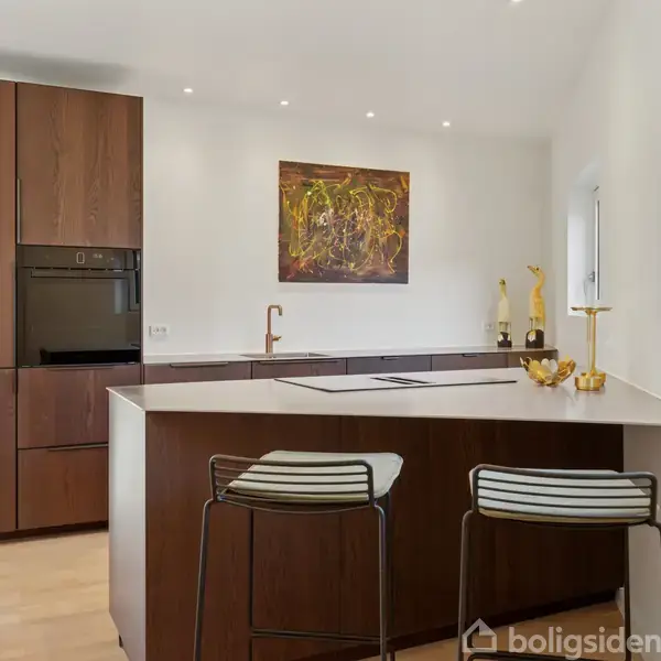 Køkkenø med to barstole står i et moderne køkken med brune skabe, indbygget ovn og abstrakt kunstværk på væggen. Lys træparketgulv og vinduer tilfører rummet lys.