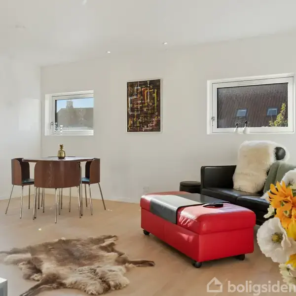 Stue med sort-rød sofa og sofapude, blomstervase foran. Spisebord med fire stole ved siden af store vinduer, hvor solskin lyser rummet. Hyggelig og moderne indretning.