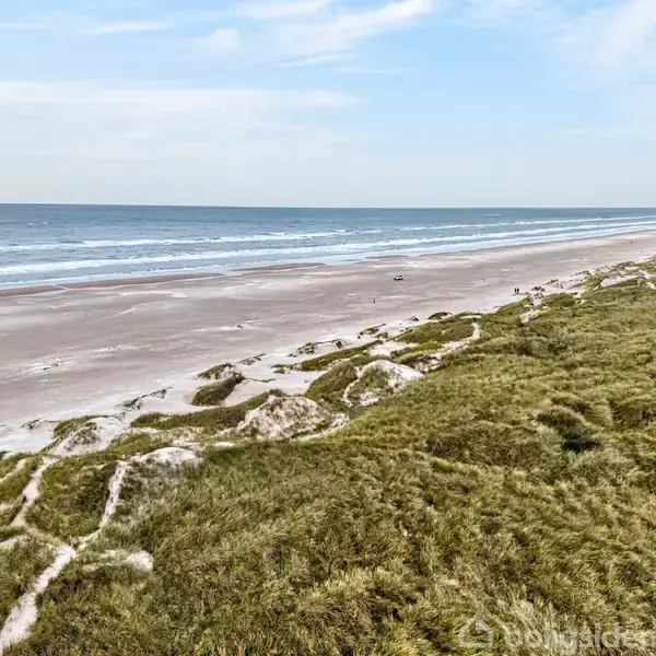 Klitter dækket af græs strækker sig langs en bred sandstrand, hvor bølger ruller ind fra havet under en klar, blå himmel.