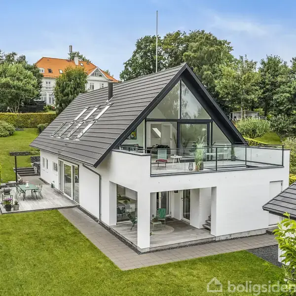 En moderne, hvid villa med skråt tag har store glaspartier og en rummelig terrasse. Huset ligger i en velplejet have med græsplæne og omkranses af buske og træer.