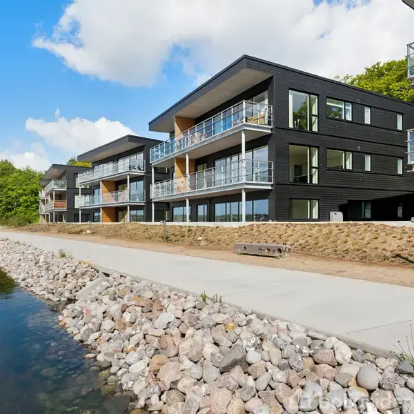 Sort moderne lejlighedskompleks med glasbalkoner, der står ved siden af en sti langs en stenet kystlinje. Flere bygninger ses i baggrunden under en blå himmel med skyer.