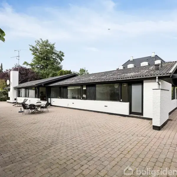 En hvid murstensvilla står stille på en flisebelagt terrasse. I forgrunden er der havemøbler. Omgivet af træer i et boligområde. Ingen tekst er synlig bortset fra "Boligsiden".