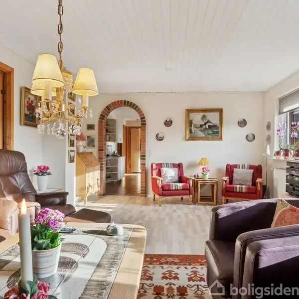 Et hyggeligt, velindrettet stueområde med brune lænestole og spisebord. Stearinlys og blomster pryder bordet. Lysekrone hænger fra loftet. I baggrunden ses et køkken gennem en buegang.