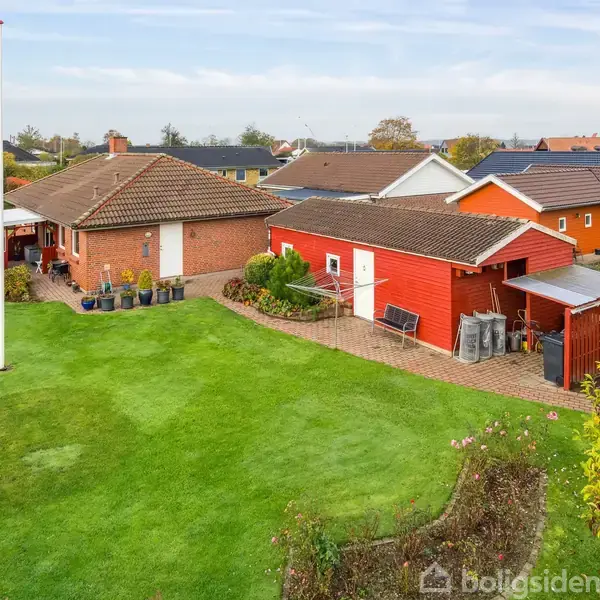 Et rødt skur med sorte døre står ved siden af en rød murstensvilla, omgivet af en stor, velplejet græsplæne og hække i et villakvarter.