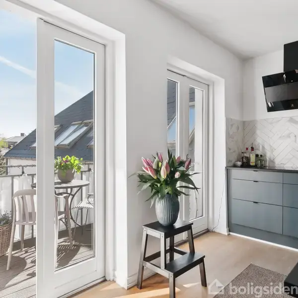 En lys lejlighedskøkken med en stor altandør, der fører ud til en balkon med planter og stole. På en taburet står en vase med blomster.